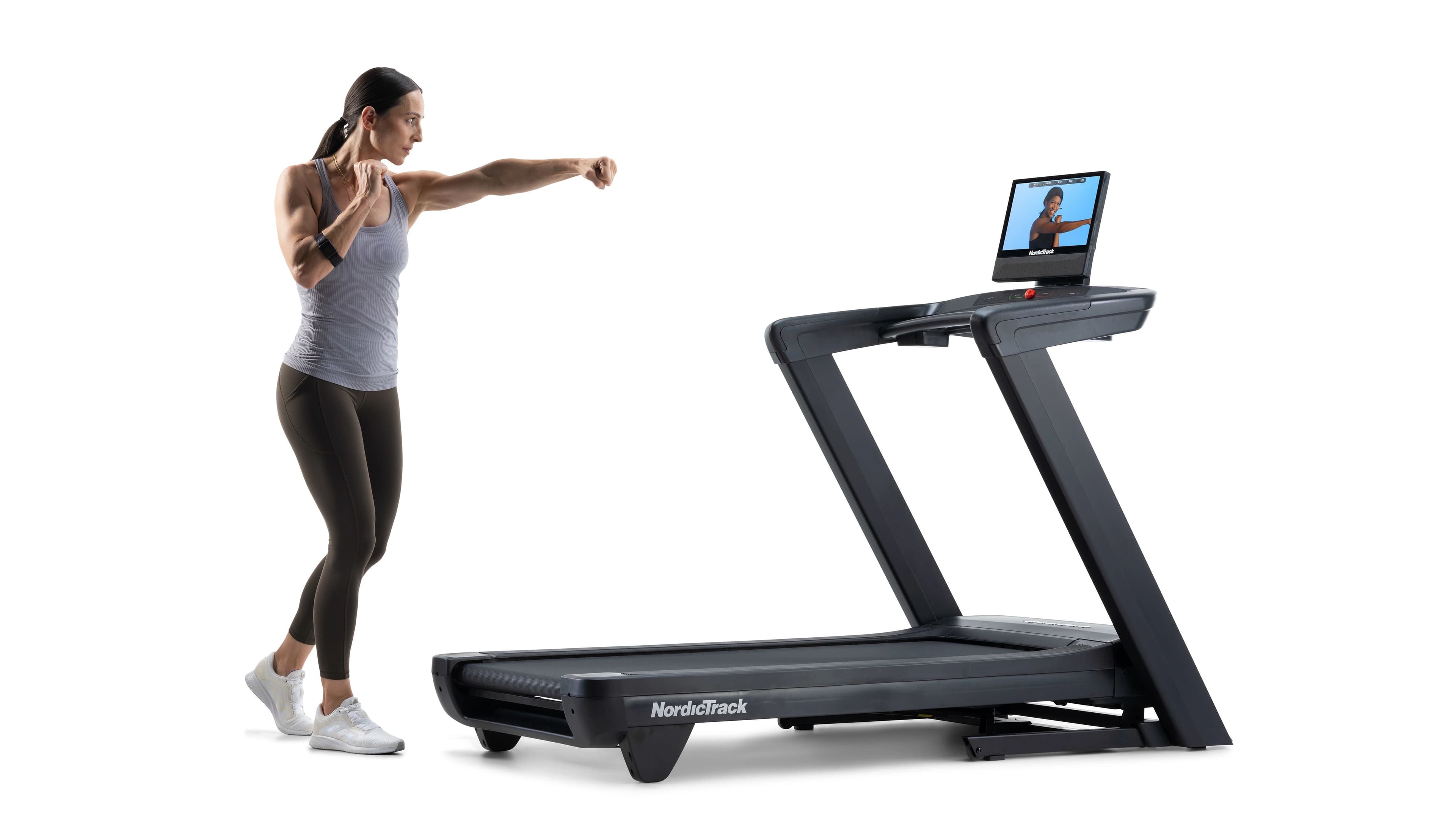 Woman in athletic wear performing a boxing punch next to a NordicTrack Commercial 1750 Treadmill with an iFIT workout displayed on the 16-inch touchscreen, white studio background