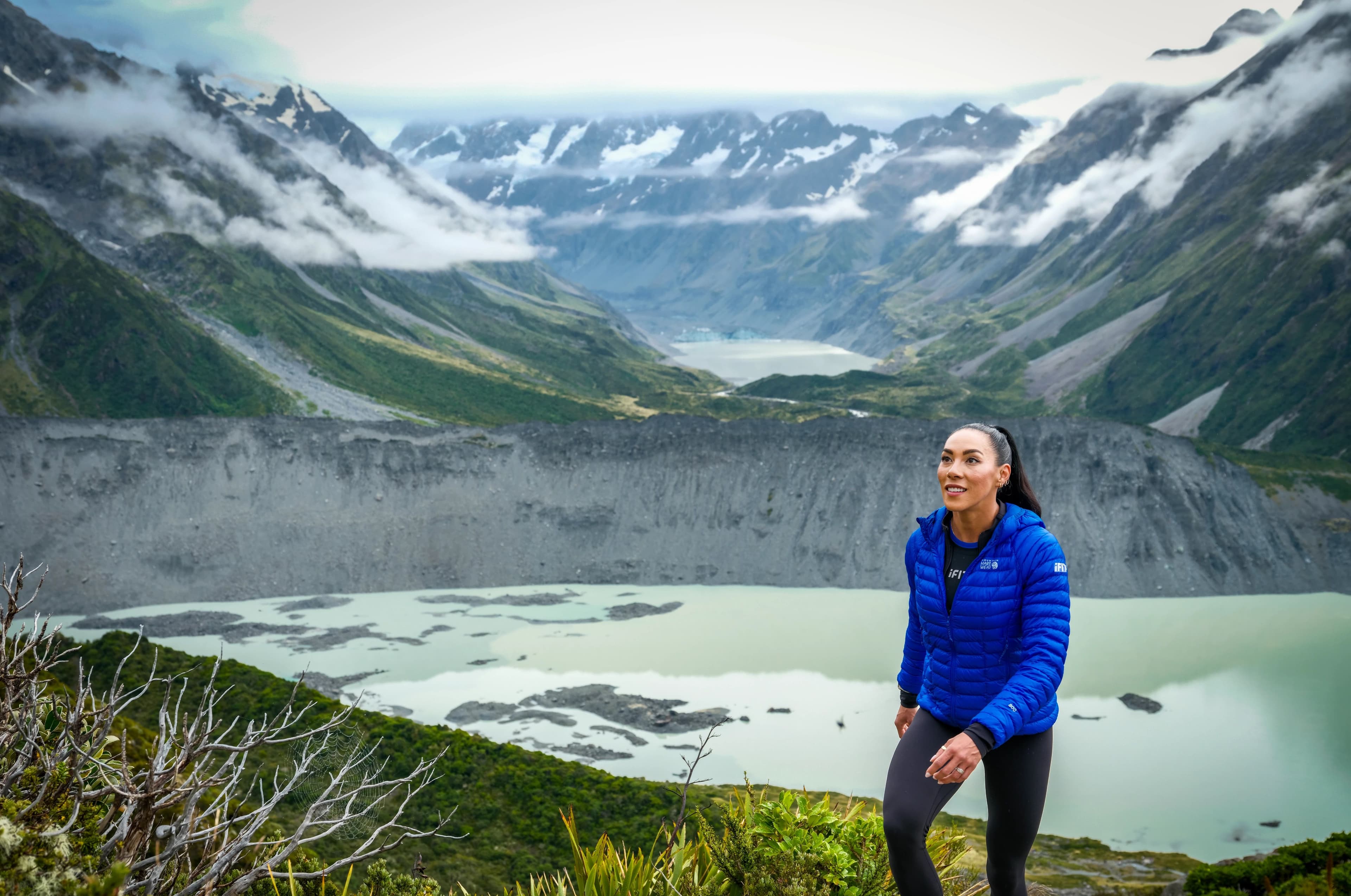 iFIT trainer Hannah Eden pausing on a hillside overlooking a glacial lake and snow-capped mountain valley in New Zealand, wearing a blue iFIT jacket