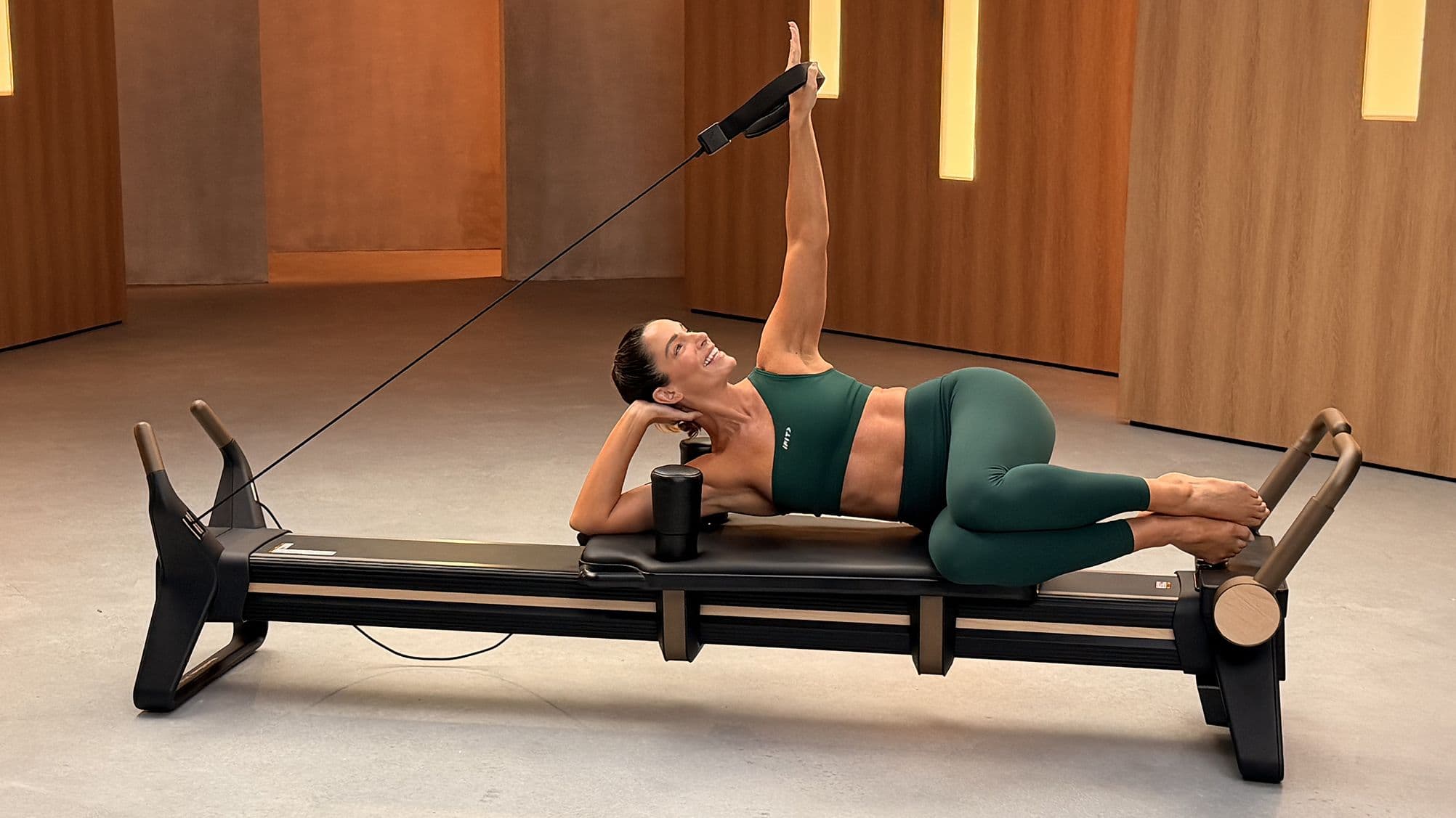 La coach iFIT Jeni DelPozo réalise un exercice de Pilates reformer en position allongée sur le côté sur le NordicTrack Ultra 1 Reformer, un bras tendu au-dessus de la tête.