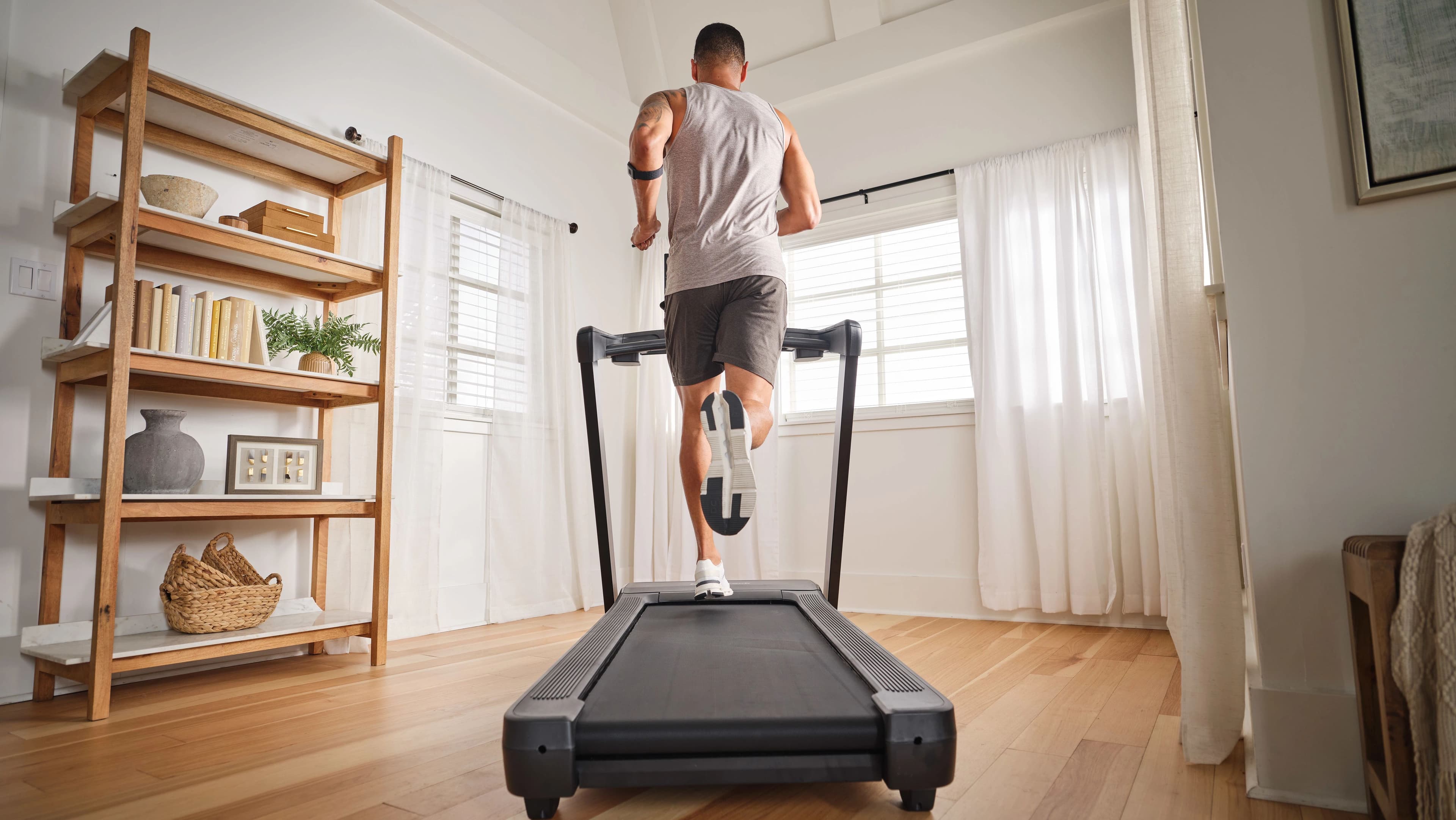 Man running on the NordicTrack T Series 10 treadmill in a bright home living room, wearing an arm-mounted heart rate monitor.