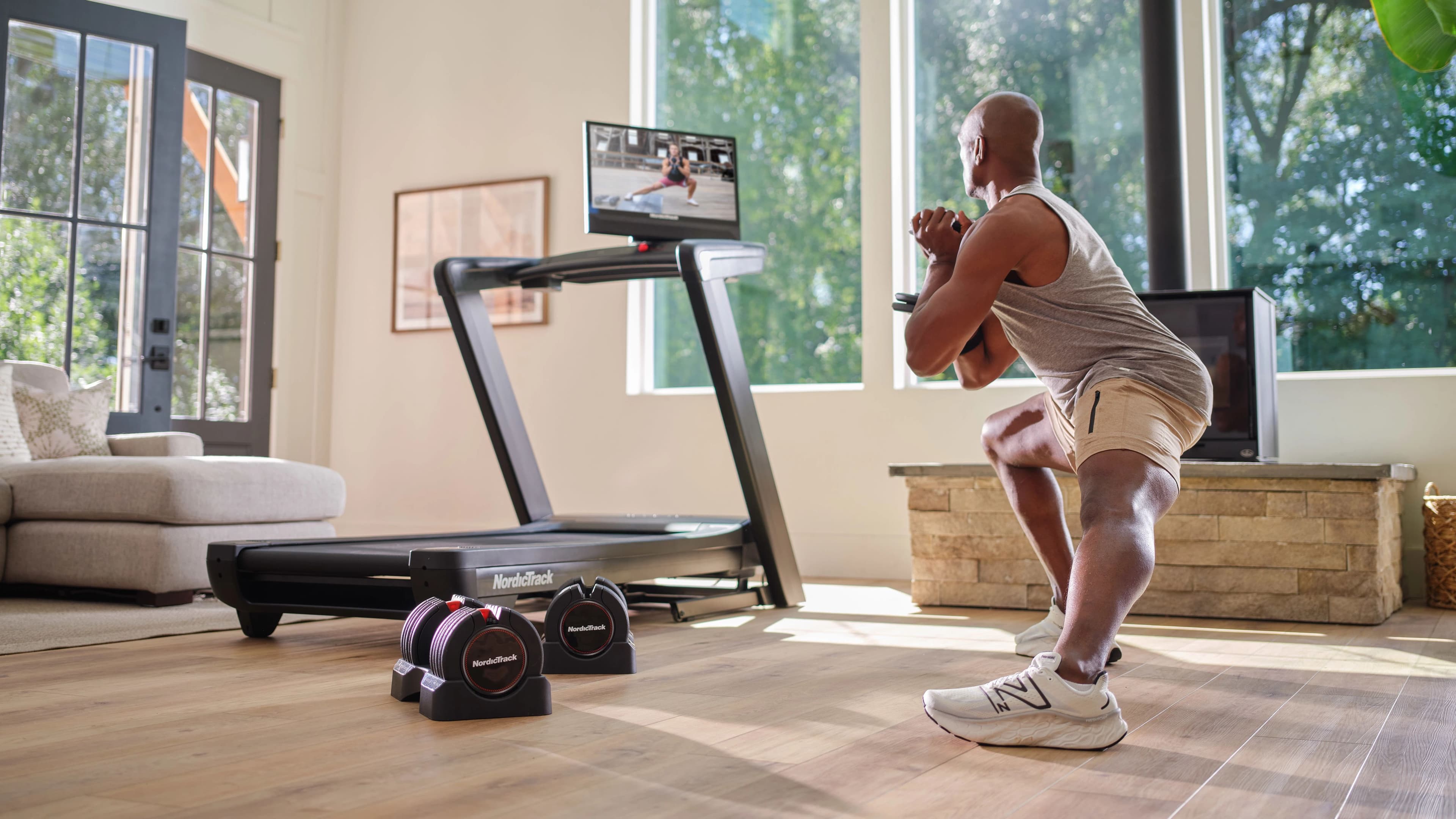 Man doing a bodyweight squat beside the NordicTrack Commercial 2450 Treadmill and adjustable dumbbells, with an iFIT trainer workout on screen