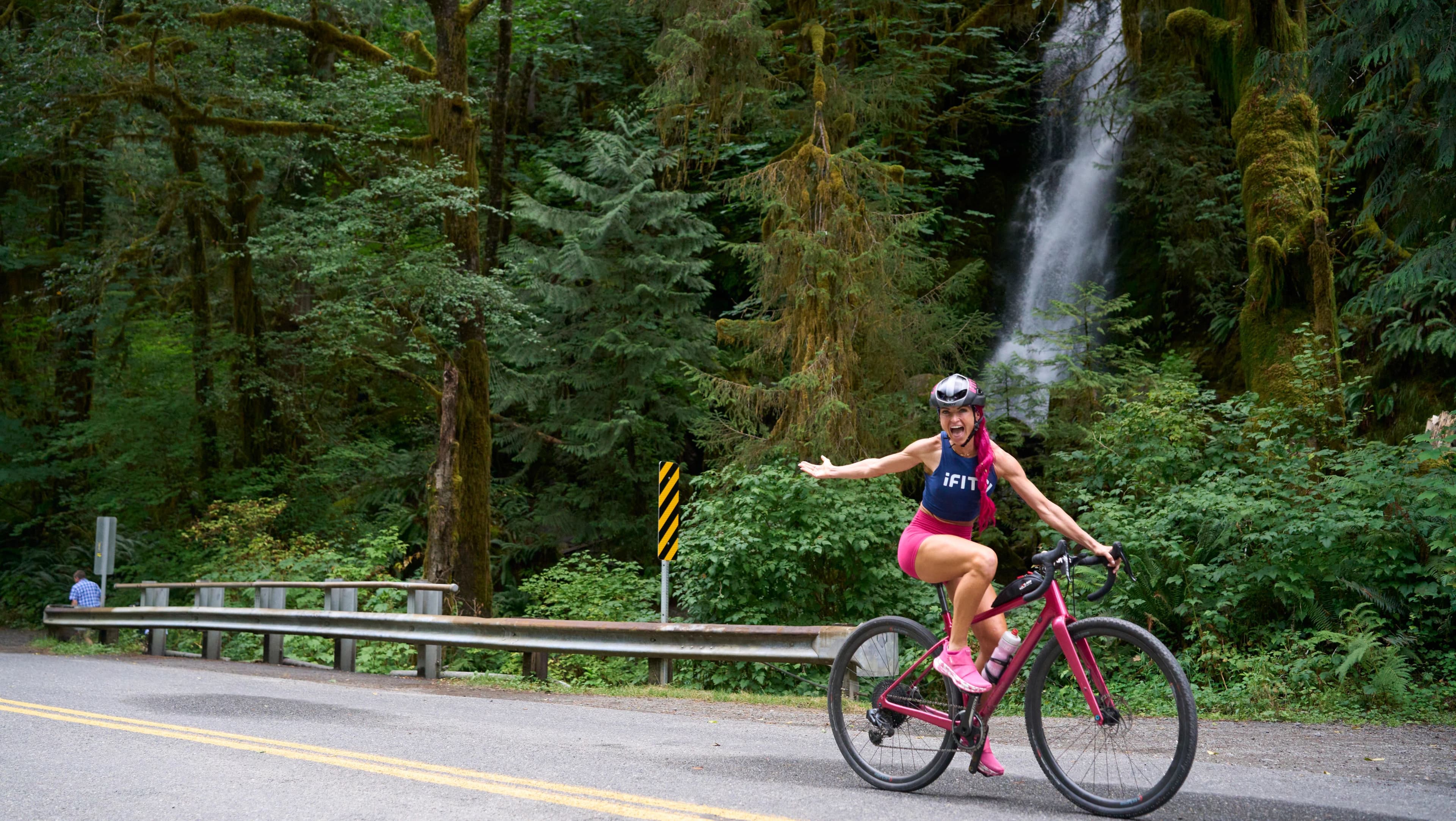 iFIT trainer Ashley Paulson riding a road bike past a waterfall on a forest road during the Washington National Parks Power and Incline Cycling Series