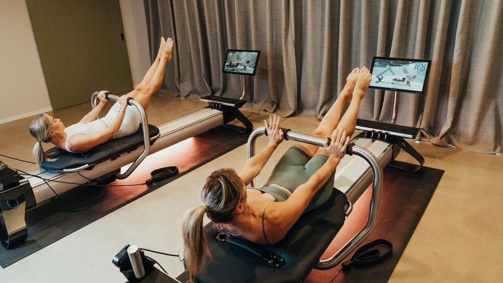 Two individuals performing controlled leg raise exercises on Pilates reformers, emphasizing core stability and guided resistance training.