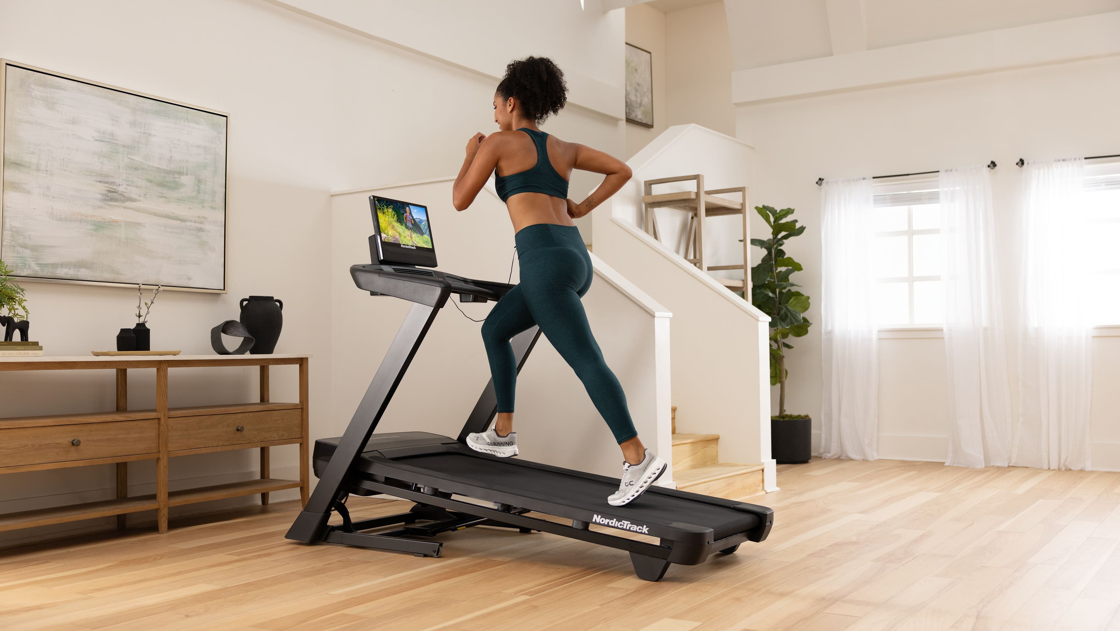 Woman running on the NordicTrack T Series 16 treadmill in a home gym, following an iFIT trail workout on the built-in touchscreen.